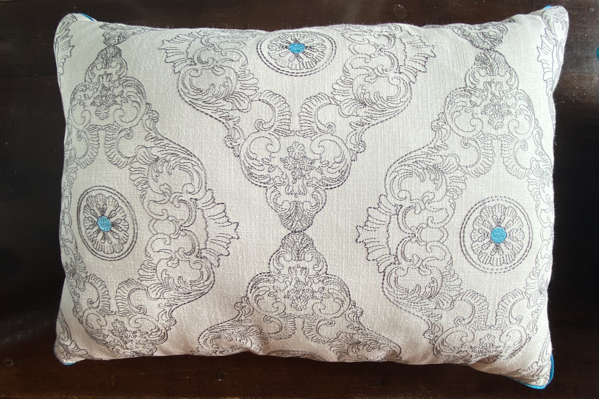 Cuscino in cotone ricamato con sfondo bianco, dettagli turchesi e bordi in cotone turchese, esempio di arredamento sostenibile e artigianale.