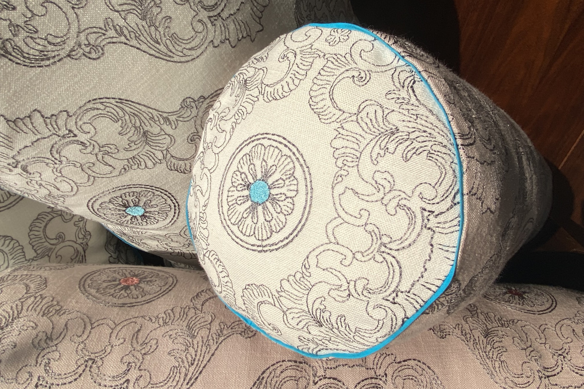 Cuscini cilindrici in cotone rosa chiaro con ricami grigi e rosa pesca, bordi turchesi e tessuti abbinati, perfetti per arredare la casa con un tocco di eleganza.