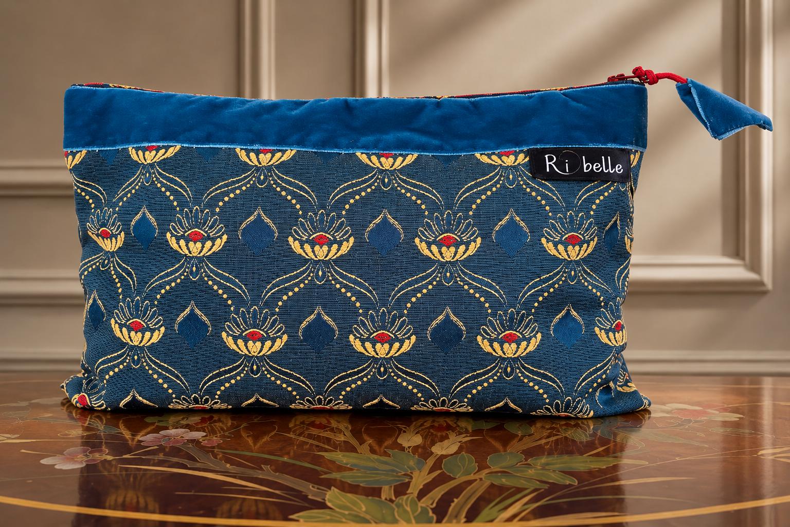Pochette artigianale in tessuto damascato blu con motivi ornamentali beige dorato e rossi, fascia in velluto blu e nappina coordinata.